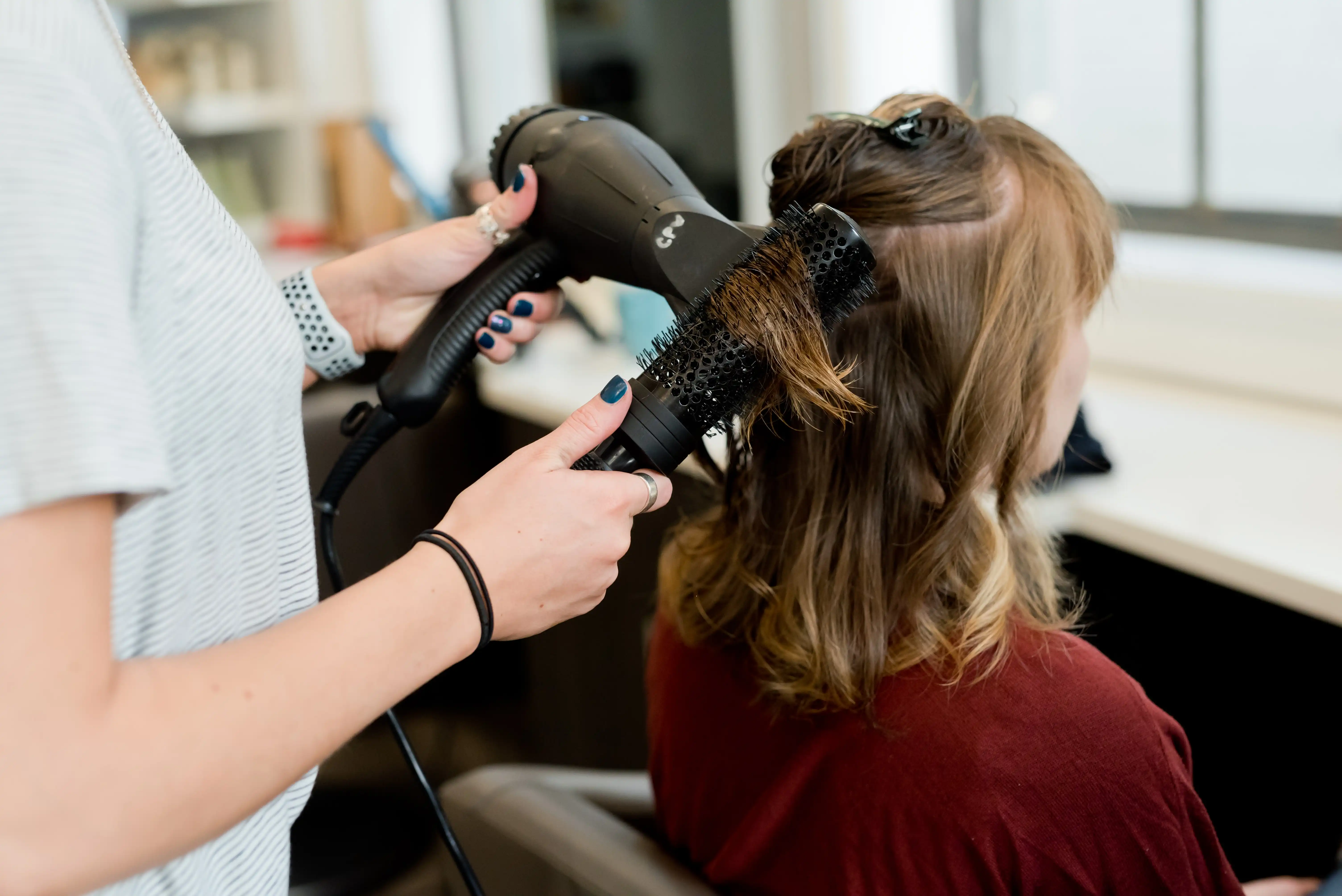 Salon hairstylist blow‑drying and brushing client’s light brown hair for smooth, voluminous style.