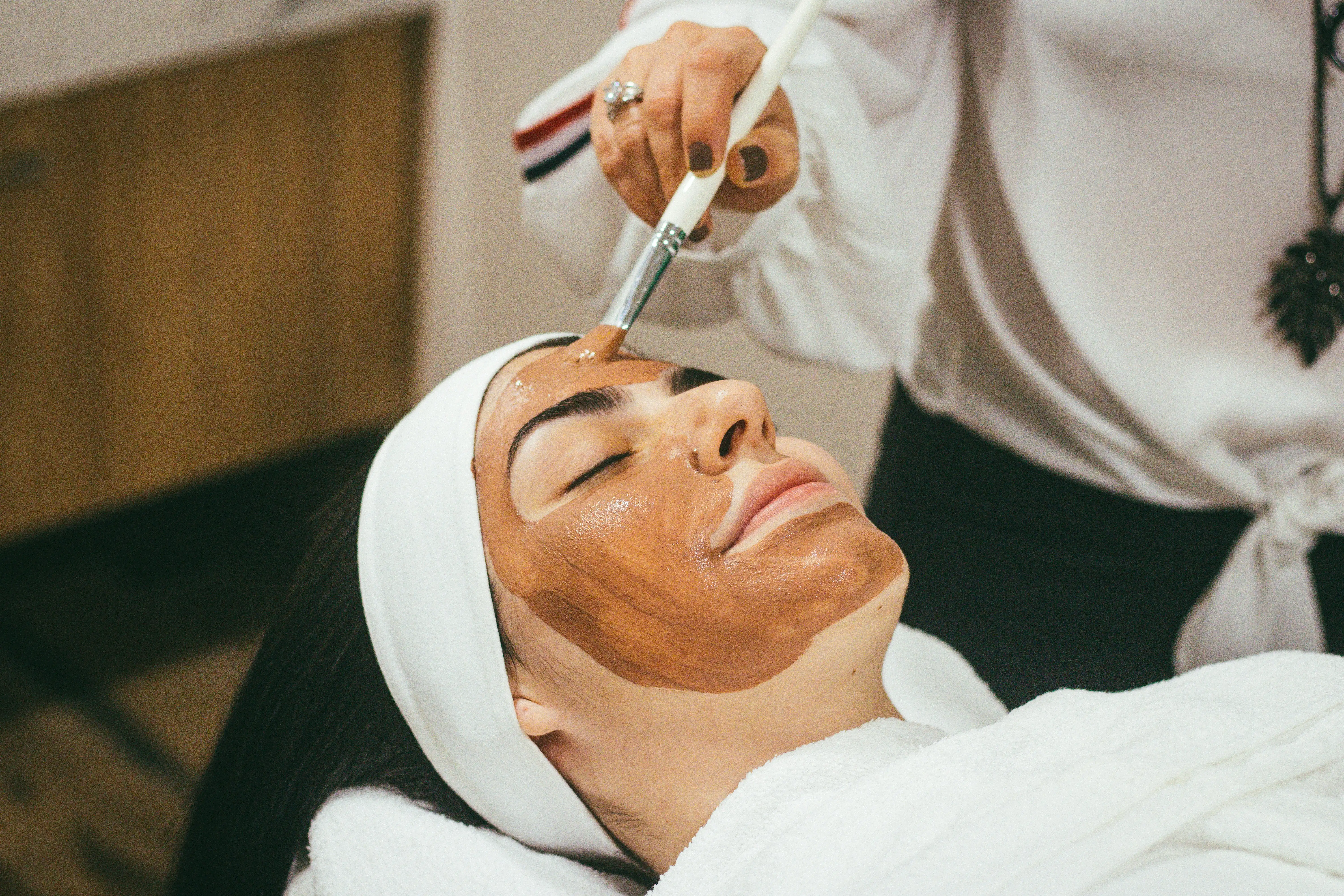 Spa facial treatment with therapist applying brown mask using brush for relaxation and skincare.